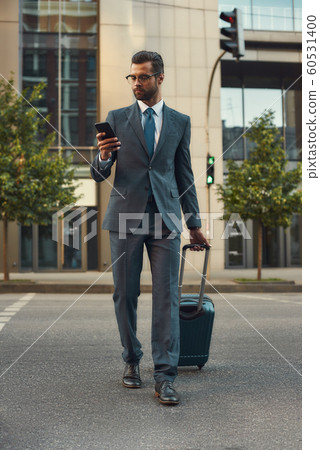Always in touch. Full length of young and handsome bearded man in suit pulling his luggage and looking at his smartphone while walking outdoors Always in touch. Full length of young and handsome bearded man in suit pulling his luggage and looking at his smartphone while walking outdoors 60531400