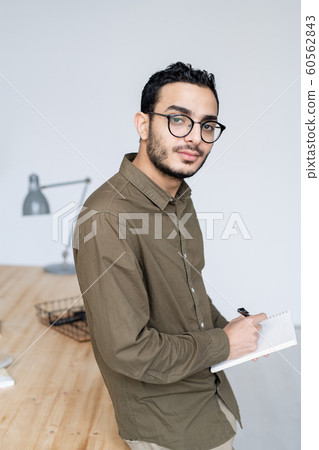Young businessman in casualwear and eyeglasses looking at you while making notes Young businessman in casualwear and eyeglasses looking at you while making notes 60562843