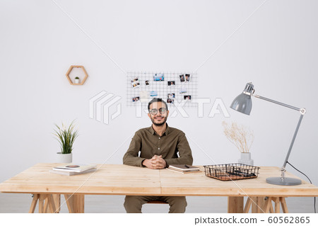 Young successful businessman in casualwear sitting by wooden table in office Young successful businessman in casualwear sitting by wooden table in office 60562865