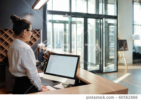 Young female receptionist standing in front of computer screen by counter Young female receptionist standing in front of computer screen by counter 60562866