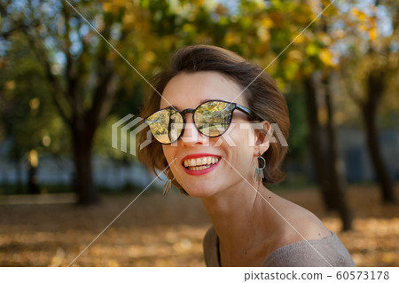 Young and beautiful girl with short hair and sunglasses with mirror surface is posing against the trees with yellow leaves background spending time in the autumn park Young and beautiful girl with short hair and sunglasses with mirror surface is posing against the trees with yellow leaves background spending time in the autumn park 60573178