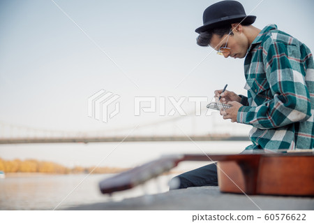 Young guy with guitar sitting near river 60576622