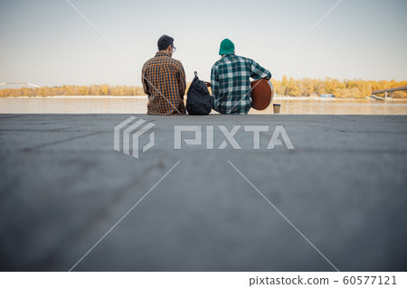 Two male friends siting with guitar near river Two male friends siting with guitar near river 60577121