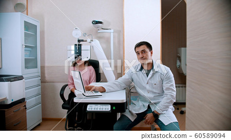Ophthalmology - young woman checking her visual acuity with an optometrist equipment - a doctor sitting near Ophthalmology - young woman checking her visual acuity with an optometrist equipment - a doctor sitting near 60589094