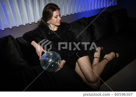 Pretty girl in black glittering dress holding disco ball while relaxing on couch Pretty girl in black glittering dress holding disco ball while relaxing on couch 60596307