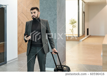 Young elegant businessman pulling suitcase while moving along hotel lounge Young elegant businessman pulling suitcase while moving along hotel lounge 60596416