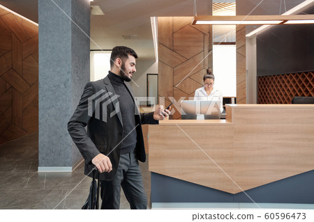 Young business traveler with suitcase scrolling in smartphone in hotel lounge Young business traveler with suitcase scrolling in smartphone in hotel lounge 60596473