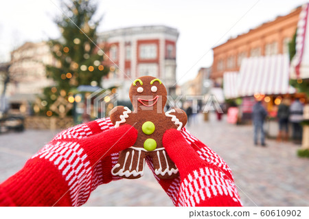 Hand in red mitten holding a smiling gingerbread man and christmas mood in blurred background. Christmas market in old town European small city. 60610902