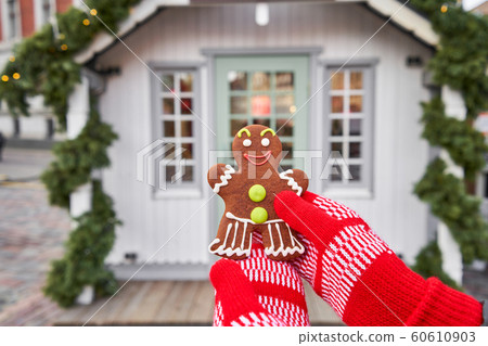 Hand in red mitten holding a smiling gingerbread man and christmas mood in blurred background. Christmas market in old town European small city. Hand in red mitten holding a smiling gingerbread man and christmas mood in blurred background. Christmas market in old town European small city. 60610903