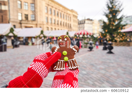 Hand in red mitten holding a smiling gingerbread man and christmas mood in blurred background. Christmas market in old town European small city. Hand in red mitten holding a smiling gingerbread man and christmas mood in blurred background. Christmas market in old town European small city. 60610912