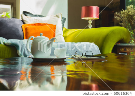 White teapot with cup and eyeglasses on table with colorful couch in background. 60616364