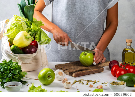 Woman cuts a green apple and cooking a healthy diet food with a cotton eco bag full of fresh vegetables on the table. Woman cuts a green apple and cooking a healthy diet food with a cotton eco bag full of fresh vegetables on the table. 60629662