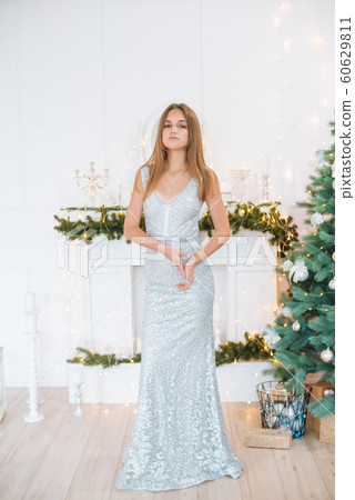 Attractive young girl with blond hair and evening make-up in an evening dress in a floor dress near a Christmas tree and a fireplace in a bright studio. New Year 2020 60629811