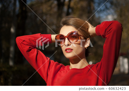 Beautiful smiling woman wearing a sunglasses and red sweater is posing in autumn park during sunny weather outdoors Beautiful smiling woman wearing a sunglasses and red sweater is posing in autumn park during sunny weather outdoors 60632423