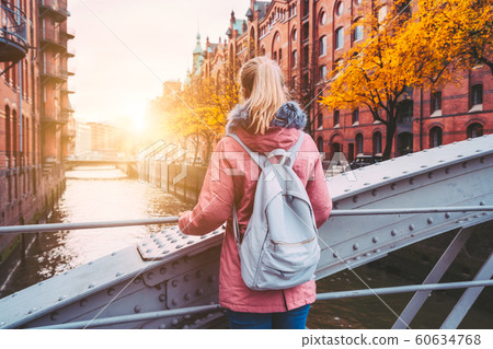 Back rear view of adult blond woman tourist with backpack enjoying autumn beautiful sunset scene on the Speicherstadt historic warehouse district in Hamburg, Germany, Europe Back rear view of adult blond woman tourist with backpack enjoying autumn beautiful sunset scene on the Speicherstadt historic warehouse district in Hamburg, Germany, Europe 60634768