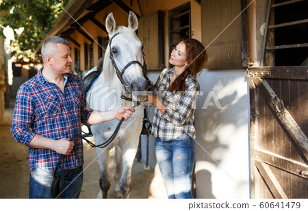 Smiling couple with white horse standing at stable outdoor Smiling couple with white horse standing at stable outdoor 60641479