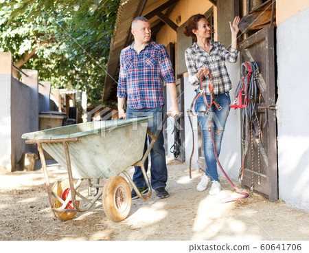 Mature positive family couple with wheelbarrow standing at horse stable 60641706