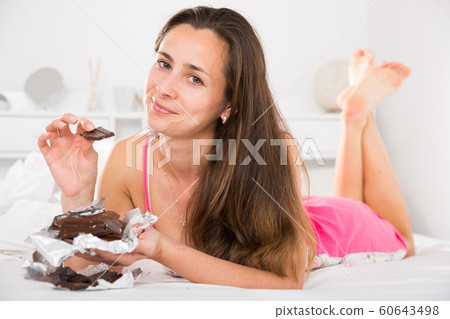 Cute female in sleepwear eating chocolate in bed in home interior Cute female in sleepwear eating chocolate in bed in home interior 60643498