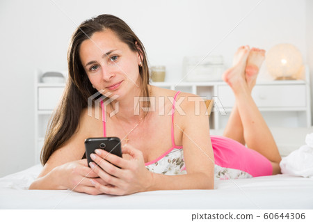 Smiling girl resting and using smartphone on bed in bedroom Smiling girl resting and using smartphone on bed in bedroom 60644306