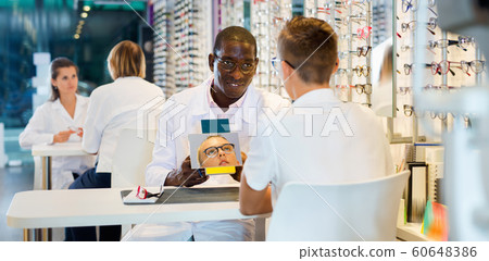 Smiling African-American ophthalmologist helping teen boy to choose glasses in optical store, showing mirror 60648386