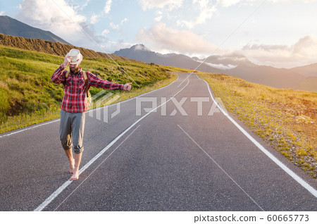 Stylish barefoot bearded male hitchhiker traveler in a hat and with a backpack walks along a suburban asphalt road in the mountains at sunset and stops the car with a hitch Stylish barefoot bearded male hitchhiker traveler in a hat and with a backpack walks along a suburban asphalt road in the mountains at sunset and stops the car with a hitch 60665773