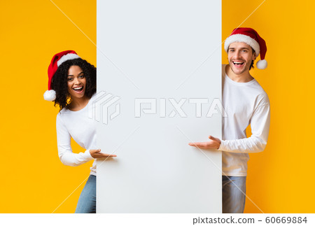 Cheerful Interracial Couple In Santa Hats Indicating At White Advertisement Board 60669884