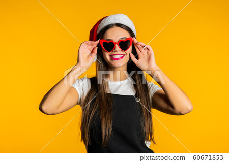 Beautiful woman with long hair in Santa hat and red heart-shaped sunglasses on studio yellow background. Beautiful woman with long hair in Santa hat and red heart-shaped sunglasses on studio yellow background. 60671853