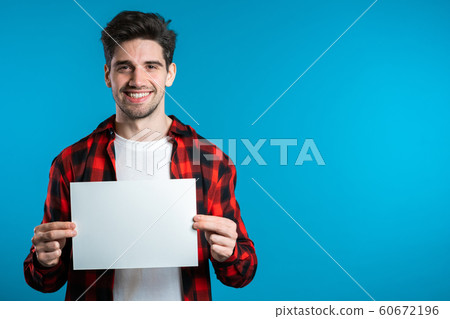 Handsome man holding white a4 paper poster. Copy space. Smiling hipster guy in red plaid shirt on blue background. Handsome man holding white a4 paper poster. Copy space. Smiling hipster guy in red plaid shirt on blue background. 60672196