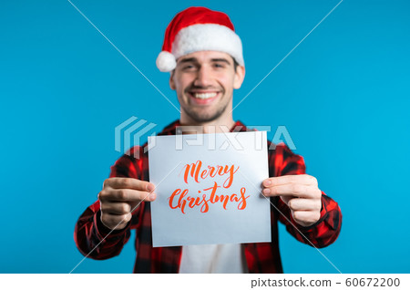 Attractive man in Santa hat with nameplate Merry Christmas. Portrait of guy on blue background. New year concept. Happy boy smiling to camera. Attractive man in Santa hat with nameplate Merry Christmas. Portrait of guy on blue background. New year concept. Happy boy smiling to camera. 60672200
