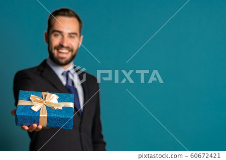 Handsome businessman in suit gives gift box and hands it to camera. He is happy, smiling. Man with beard on blue background. Positive holiday shot. Handsome businessman in suit gives gift box and hands it to camera. He is happy, smiling. Man with beard on blue background. Positive holiday shot. 60672421