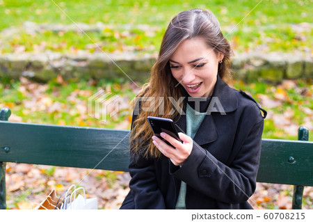 Beautiful girl sitting on a bench, brunette woman with your phone writes a message on social networks, sunny day in the park, relaxing, after work business woman 60708215