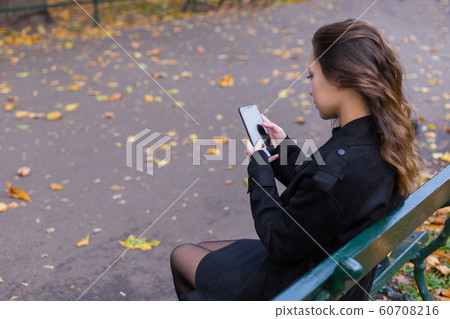 Beautiful girl sitting on a bench, woman with your phone writes a message on social networks, sunny autumn day in the park, woman relaxing after shopping. Mockup for your design 60708216