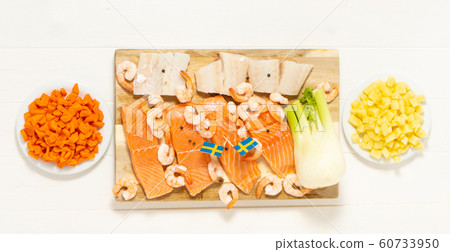 Salmon, cod, shrimps, fennel on a wooden cutting board. Potatoes and carrots in white plates. Swedish flags. Ingredients for traditional Scandinavian fish soup. Salmon, cod, shrimps, fennel on a wooden cutting board. Potatoes and carrots in white plates. Swedish flags. Ingredients for traditional Scandinavian fish soup. 60733950