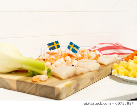 Salmon, cod, shrimps, fennel on a wooden cutting board. Potatoes and carrots in white plates. Swedish flags. Ingredients for traditional Scandinavian fish soup. Salmon, cod, shrimps, fennel on a wooden cutting board. Potatoes and carrots in white plates. Swedish flags. Ingredients for traditional Scandinavian fish soup. 60733954
