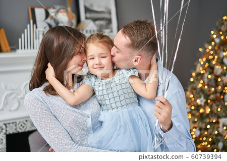 Loving family. Mom and dad hugging little daughter . Parents and baby child having fun near Christmas tree and white fireplace indoors. Merry Christmas and Happy New Year. Cheerful pretty people. 60739394