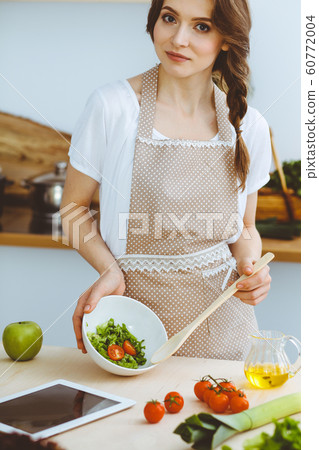 Young brunette woman cooking in kitchen. Housewife holding wooden spoon in her hand. Food and health concept Young brunette woman cooking in kitchen. Housewife holding wooden spoon in her hand. Food and health concept 60772004