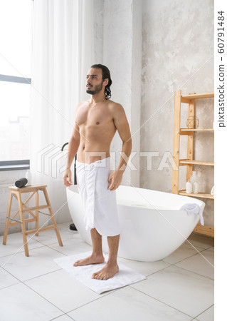 Young shirtless man with soft white towel on hips standing on small rug Young shirtless man with soft white towel on hips standing on small rug 60791484