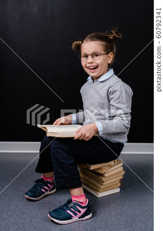 Excited adorable schoolgirl in eyeglasses and casualwear sitting on books Excited adorable schoolgirl in eyeglasses and casualwear sitting on books 60792341