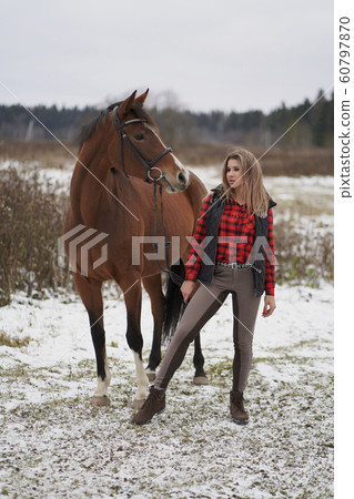 A beautiful girl in the red plaid shirt and pants rider walks with horse in early winter. space for text on white background A beautiful girl in the red plaid shirt and pants rider walks with horse in early winter. space for text on white background 60797870
