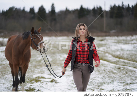 A beautiful girl in the red plaid shirt and pants rider walks with trakehner horse in early winter A beautiful girl in the red plaid shirt and pants rider walks with trakehner horse in early winter 60797873