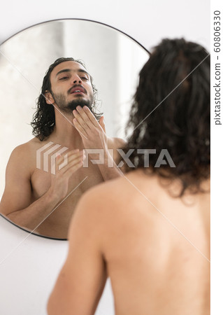 Shirtless young man looking at his beard in mirror while going to shave 60806330