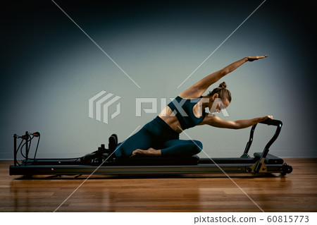 Young girl doing pilates exercises with a reformer bed. Beautiful slim fitness trainer on reformer gray background, low key, art light. Fitness concept 60815773