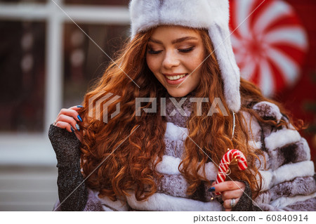 Closeup portrait of young beautiful redhead curly smiling girl is wearing hat with traditional xmas lollipop. Festive red Christmas house on background. Holiday concept. 60840914