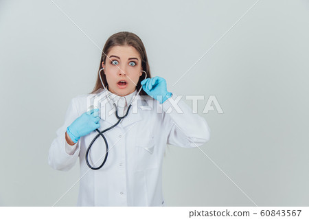 Pretty young medicine assistant standing with stethoscope, looking as if she was deafened. 60843567