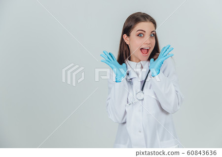 Funny female intern standing in medical uniform, raising her hands, looking at the camera with surprise and satisfaction on her face, mouth and eyes widely opened. 60843636