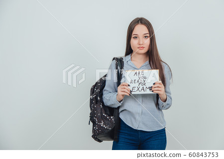 Pretty young asian girl standing, holding a sheet of paper with the prase ready for exam written on it. Backpack slung over her shoulder. Pretty young asian girl standing, holding a sheet of paper with the prase ready for exam written on it. Backpack slung over her shoulder. 60843753