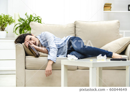 Young drunk woman with empty bottle in her hand is lying on the sofa. Woman feels headache. Young drunk woman with empty bottle in her hand is lying on the sofa. Woman feels headache. 60844649