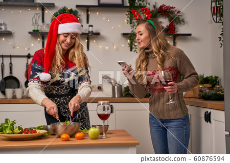 Blonde woman in santa hat prepares food and girl in Christmas deer horns with wine glass standing at kitchen. 60876594