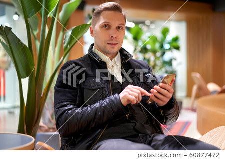 Male in leather jacket with smartphone looking at camera while sitting on chair in store against green plant Male in leather jacket with smartphone looking at camera while sitting on chair in store against green plant 60877472