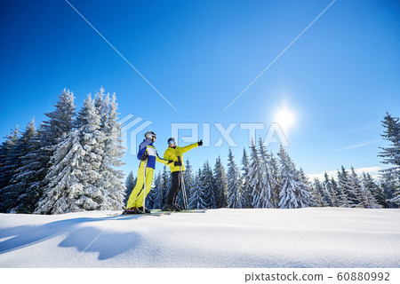 Couple of skiers on skis on hill at ski resort. Recreational activities in mountains concept. Low angle full-length shot Couple of skiers on skis on hill at ski resort. Recreational activities in mountains concept. Low angle full-length shot 60880992
